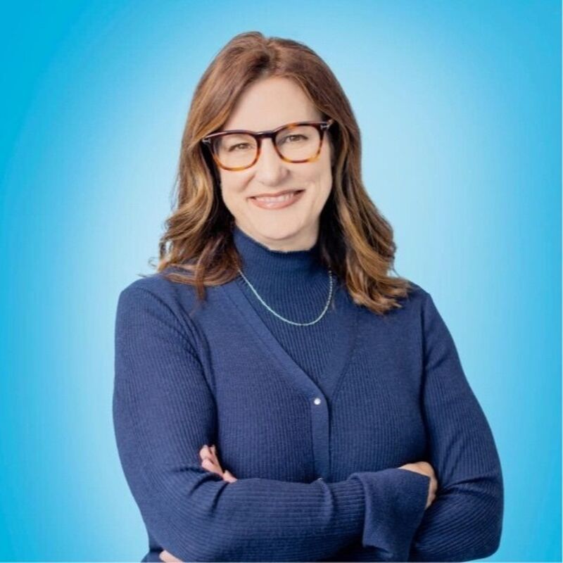 The image shows a woman with shoulder-length brown hair, wearing glasses and a navy blue sweater. She has her arms crossed and is smiling slightly. She is also wearing a thin silver necklace. The background is a gradient of light blue, creating a professional and approachable atmosphere. The lighting is soft and even, highlighting her features.